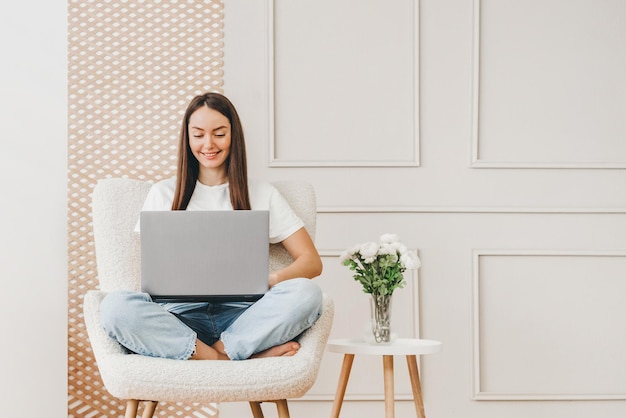 Girl freelancer sits in a chair and works with a laptop at home on the background of a white wall