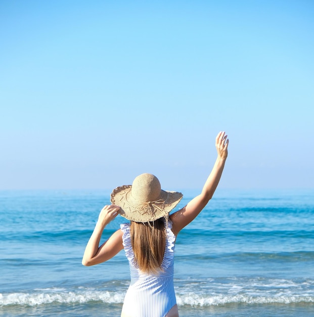 Girl from behind in blue swimsuit and straw hat with blue ocean and blue sky on background