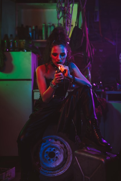 Girl in a futuristic cyberpunk costume smokes a cigarette with a neon light in the garage. Steampunk style. Futuristic post-apocalyptic style