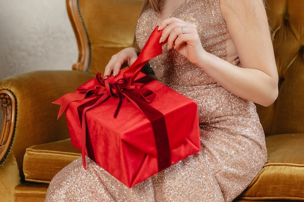 A girl in a golden dress opens a red gift while sitting on a sofa Girl's hands close up with bow and box