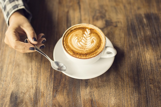 Girl hand keeping a teaspoon and cup of cappuccino on a wooden table