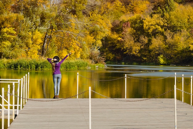 Girl in the hat is standing on the pier Hands up Autumn sunny day Back view