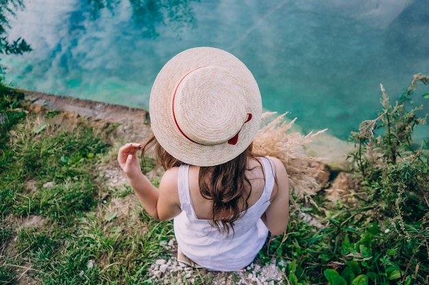 Girl in a hat sits with his back on a background of blue lake. Girl on a summer day near the wellspring. Girl in nature.