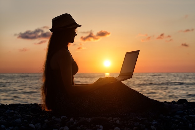 Girl in a hat sitting and working at his laptop against the sea at sunset