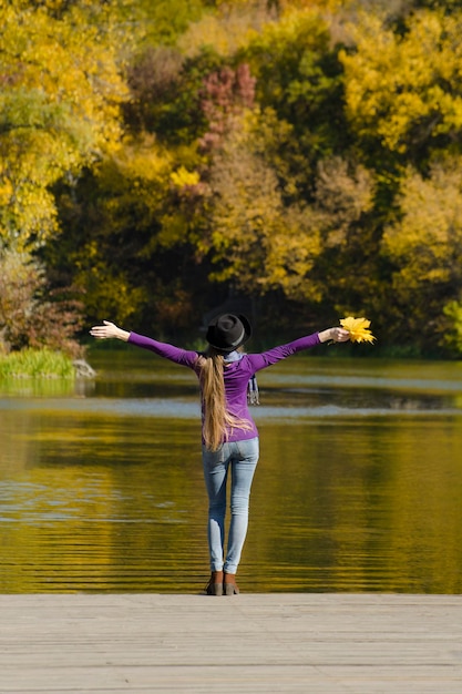 Girl in hat standing on the pier with open arms Autumn sunny day Back view