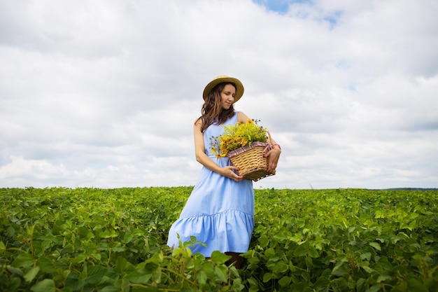 Girl in a hat stands on a green field with a basket of flowers