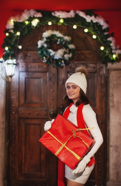 Girl in hat white dress holding red gift smiling looking at the camera Christmas or new year