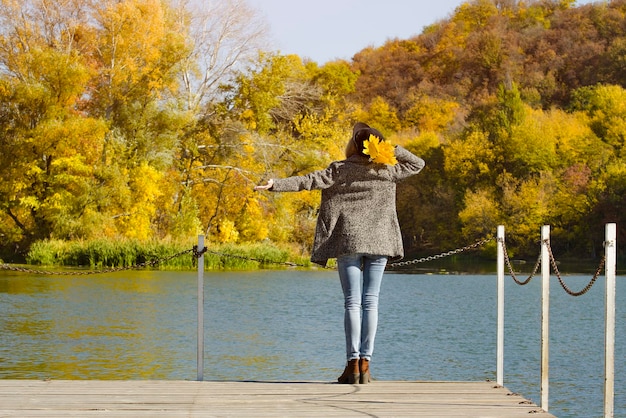 Girl in a hat with leaves in hands standing on the dock Autumn sunny Back view