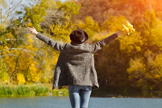 Girl in a hat with leaves in hands standing on the dock Autumn sunny Back view