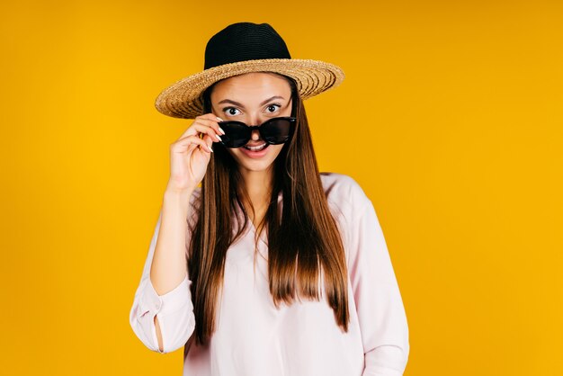 The girl in the hat, with surprise, lowered her glasses to her nose with her hand and looks over them. studio yellow