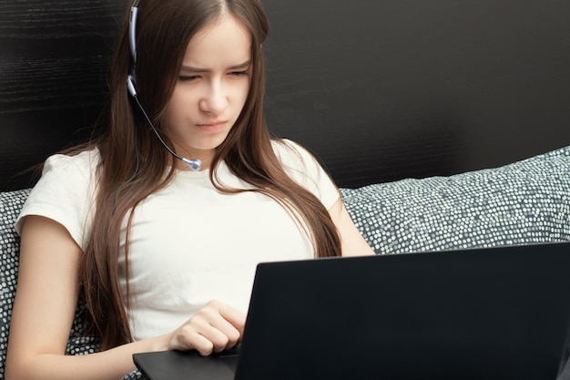 Girl in headphones with a microphone in front of a laptop lies in bed