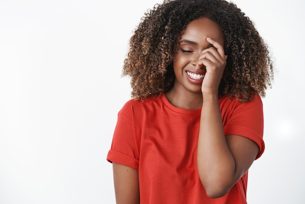 Girl hearing hilarious jokes laughing and having fun close eyes from joy and happiness touching face shy smiling broadly having good mood posing in red casual tshirt over white background