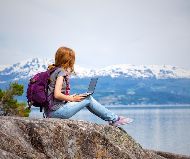 Girl hikerl with a laptop sitting on a rock on a background of mountains and lakes