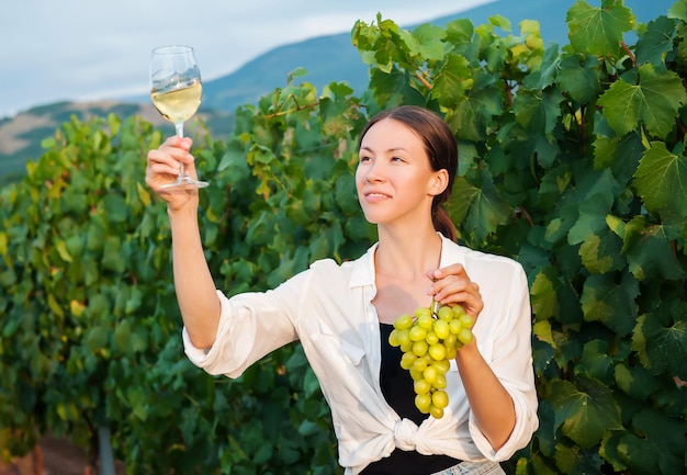 Girl holding a glass of white wine and a bunch of grapes against the backdrop of a vineyard