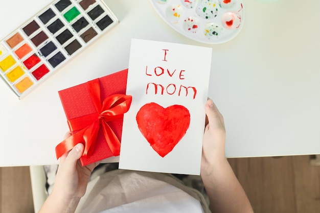 Girl holding in hands a red gift box and handmade greeting card for mom