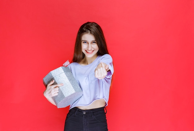 Girl holding a silver gift box and asking the person to come nearby to receive it. 