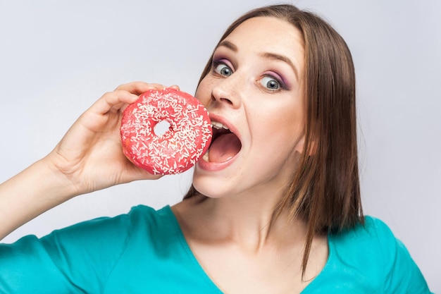 Girl holding and trying to bite big pink donut with open mouth and big eyes in grey background