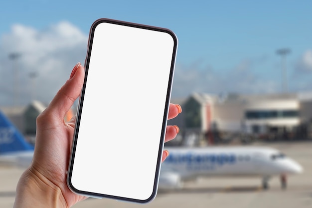 Girl holds a mock-up of a smartphone with a white screen close-up. Phone on the background of the airport with an airplane.