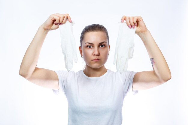 The girl holds white medical gloves in her hands. Protection against germs and the virus. She is in a white T-shirt on a white background.