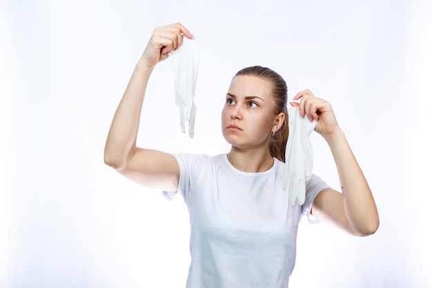 The girl holds white medical gloves in her hands. Protection against germs and the virus. She is in a white T-shirt on a white background.