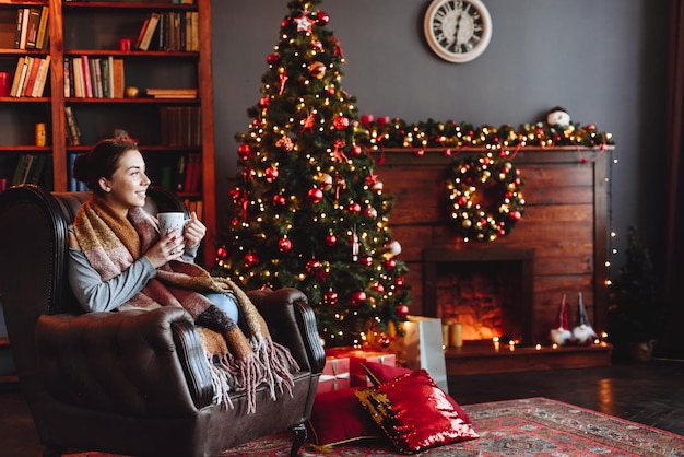 Girl at Home by Christmas tree in Warm Scarf Looks out Window Woman Holding Cup of Aromatic Drink