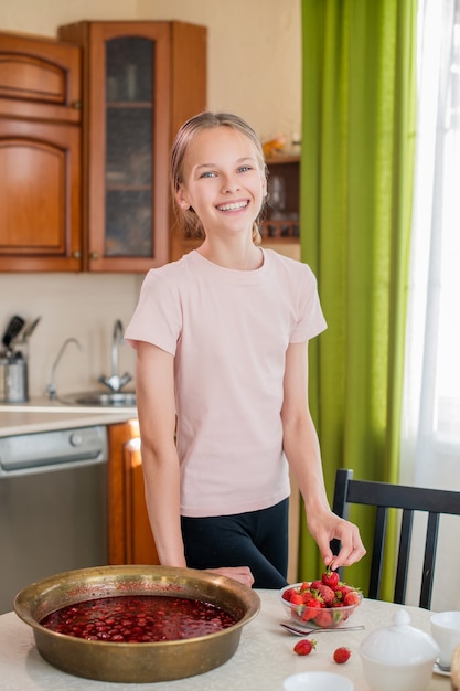 A girl at home in the kitchen stands near the table, eats strawberries, prepares strawberry jam, smiles with a perfect smile