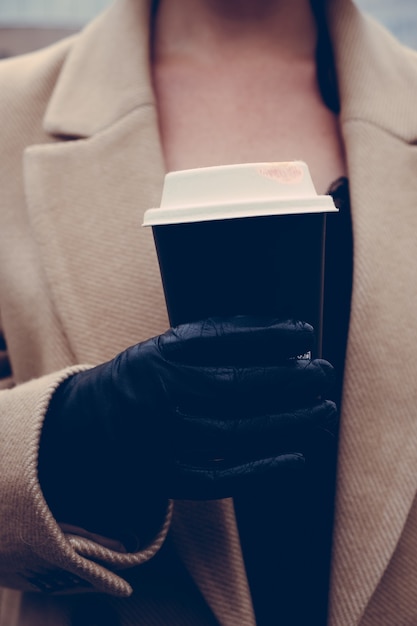 The girl is holding a paper cup with coffee in her hand, outside. Front view