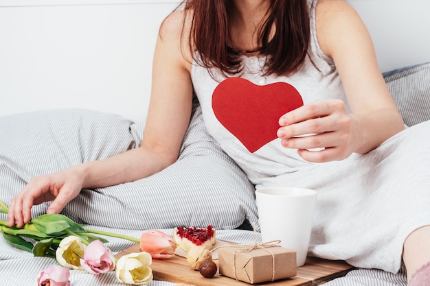 The girl is holding a red heart. Congratulations on Valentine's Day on February 14. Breakfast in bed and a gift.