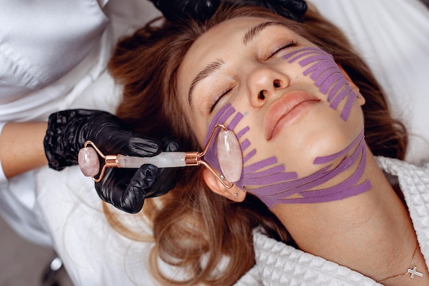 The girl is lying on the couch in the cosmetologist's office on the procedure of taping with the effect of rejuvenation receiving a massage roller