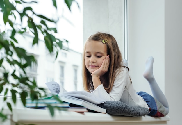 The girl is  lying on the window sill and reading a book.