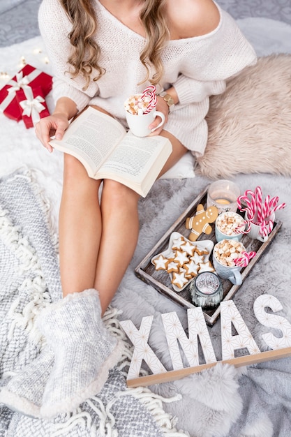 The girl is sitting in a christmas atmosphere, drinking a hot drink and reading a book.