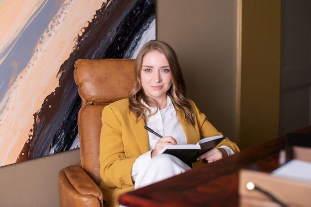 The girl is sitting at the office desk in an armchair and making notes in a notebook smiling