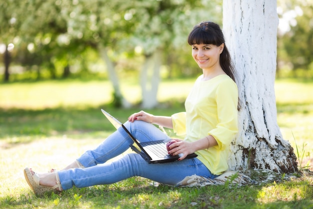 A girl is sitting with a laptop near a tree.