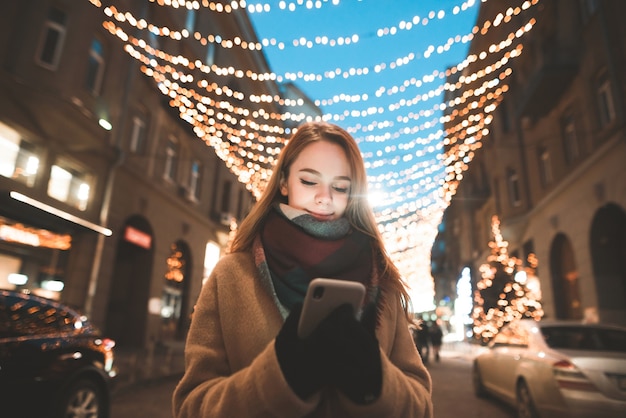 Girl is in the warm clothes standing on the street and using a smartphone, background of street lights