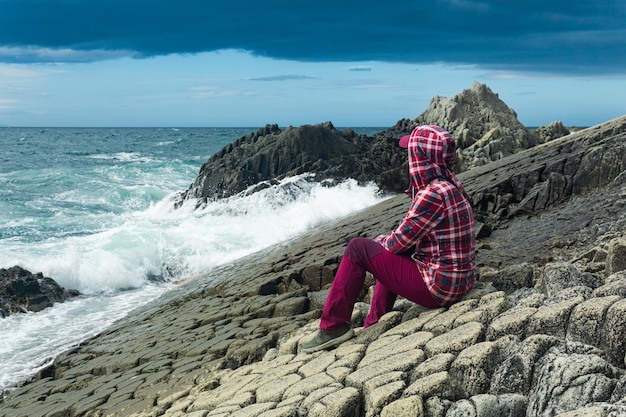 Girl in a jacket with a hood sits on the shore of a stormy sea