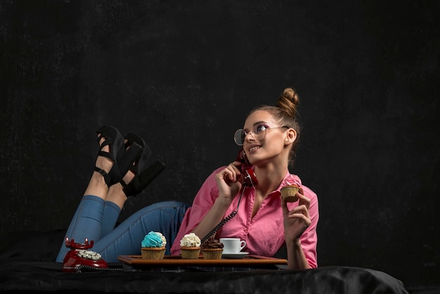 Girl lies on the bed with retro phone handset and cupcake in her hands. Portrait of stylish teenage girl on bed on a dark background.