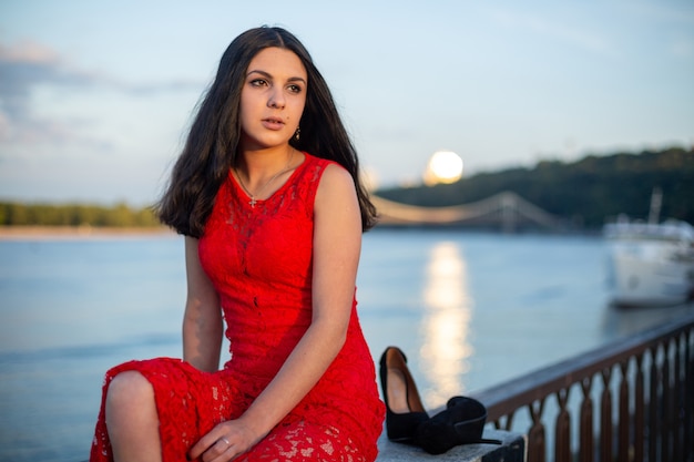 A girl in a long red dress is sitting on the fence of the river embankment.