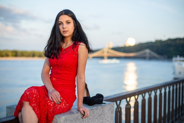 A girl in a long red dress is sitting on the fence of the river embankment.