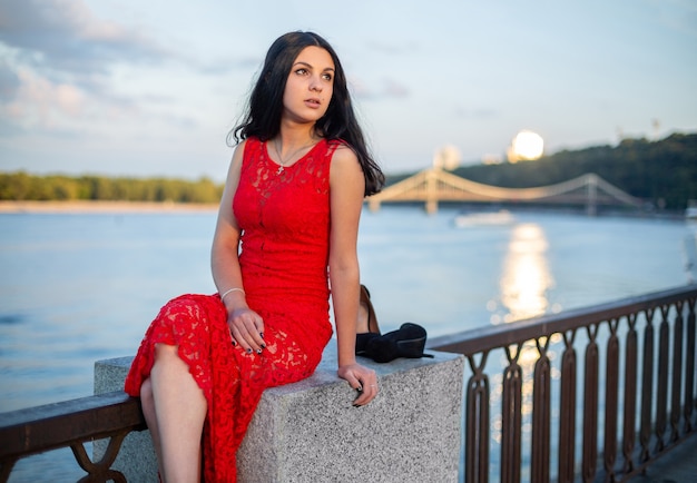 A girl in a long red dress is sitting on the fence of the river embankment.