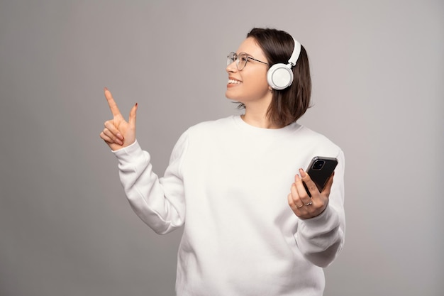 Girl looking up and wearing headphones is pointing aside and holding phone