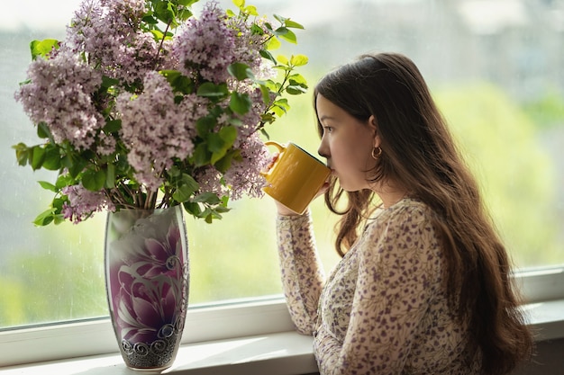 girl looks out the window and drinks tea or coffee from a yellow mug next on the windowsill there is a vase with a bouquet of lilacs