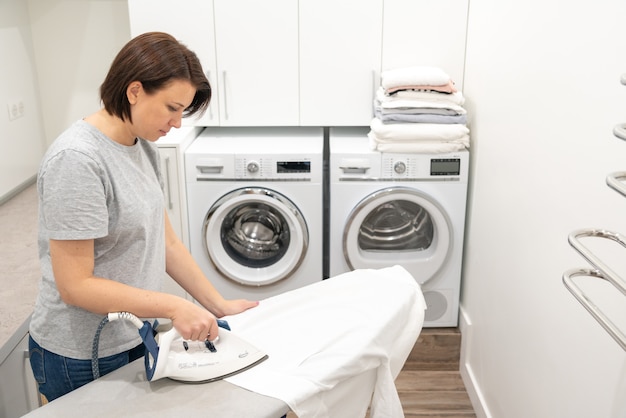 Girl in loundry room ironing white shirt on board with washing machine 