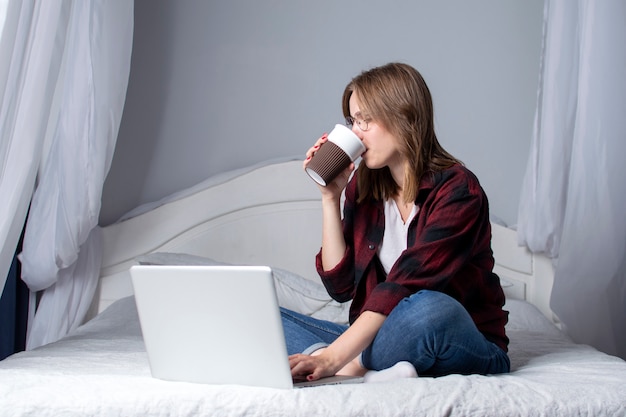 Girl lying in bed with a laptop and drinking a coffee