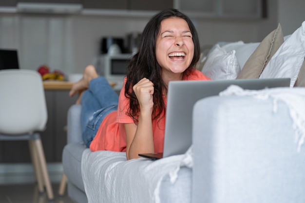 Girl lying on the couch rejoices looking at the laptop. Smiles, good mood, emotion of joy.