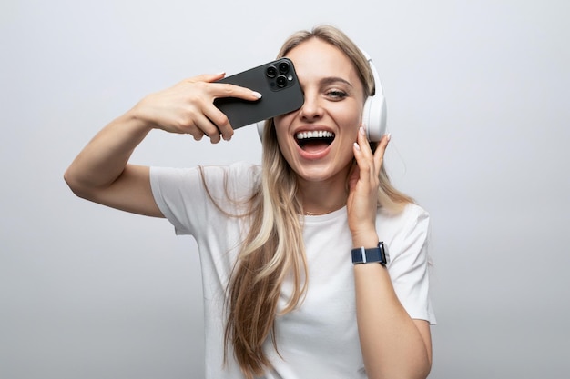 Girl makes a face in wireless headphones with a smartphone in her hands on a white background