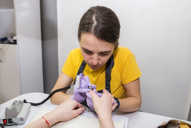 Girl master makes a manicure to a client using a milling machine in a nail salon.