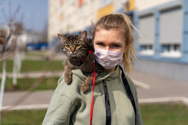 A girl in a medical mask is walking with a cat along the street near the house