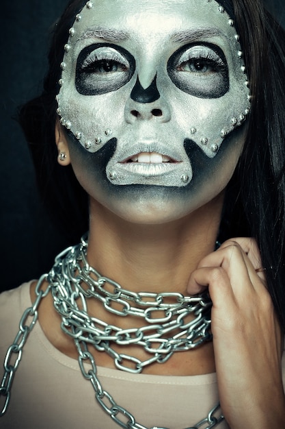 Girl model wearing chains with silver mask skull paint on dark background