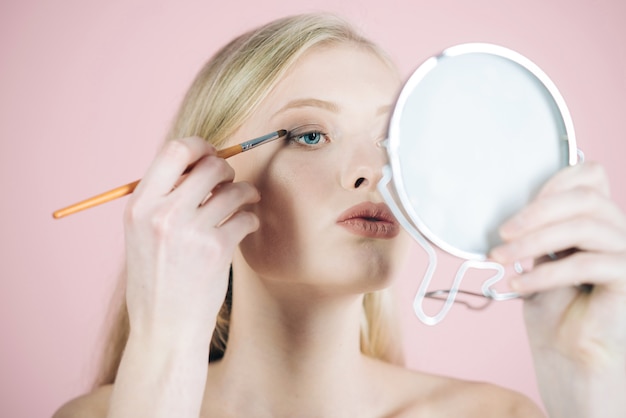 Girl model with makeup brush near face. Skin care products. Woman with blue eyes applying makeup using makeup brush and look in mirror. Light make up. Natural beauty. Skincare facial treatment concept