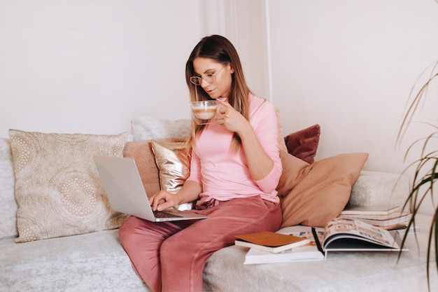 A girl in the morning in pajamas at home working on a laptop with drinking coffee, a girl self-isolated at home and resting on the couch and watching a laptop.Household chores.